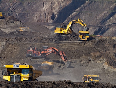 Heavy machinery operates on a large-scale mining site. Excavators and dump trucks are moving earth and materials, surrounded by rocky terrain and steep slopes. The equipment is primarily yellow and orange, indicating industrial use, and work is being actively carried out to extract resources.