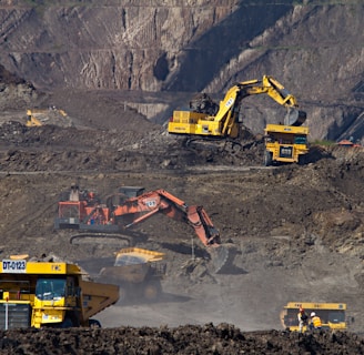 Heavy machinery operates on a large-scale mining site. Excavators and dump trucks are moving earth and materials, surrounded by rocky terrain and steep slopes. The equipment is primarily yellow and orange, indicating industrial use, and work is being actively carried out to extract resources.