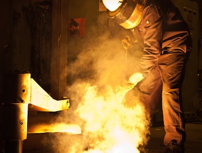 A person wearing protective gear is working in an industrial setting with molten metal creating a bright glow. The surroundings are dimly lit, emphasizing the intense light and heat coming from the metal.