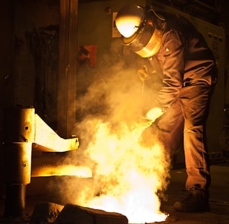 A person wearing protective gear is working in an industrial setting with molten metal creating a bright glow. The surroundings are dimly lit, emphasizing the intense light and heat coming from the metal.