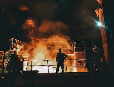 Workers stand near a large industrial furnace with bright flames and heavy smoke, suggesting a metal or steel manufacturing process. The lighting is dim with an orange glow illuminating the scene, enhancing the industrial atmosphere.