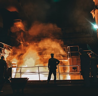 Workers stand near a large industrial furnace with bright flames and heavy smoke, suggesting a metal or steel manufacturing process. The lighting is dim with an orange glow illuminating the scene, enhancing the industrial atmosphere.