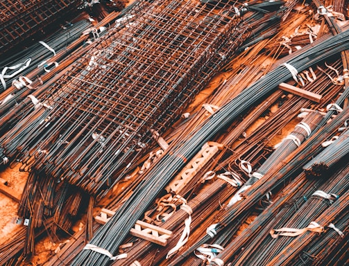 A collection of metal rods and bars are organized on the ground, tied together with what appear to be white straps. Some of these metal pieces form grid-like structures, likely reinforcing wire meshes. The setting conveys a scene typical of a construction site, where materials are being prepared or stored for use.