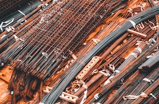 A collection of metal rods and bars are organized on the ground, tied together with what appear to be white straps. Some of these metal pieces form grid-like structures, likely reinforcing wire meshes. The setting conveys a scene typical of a construction site, where materials are being prepared or stored for use.