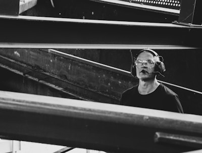 A man wearing protective eyewear and earmuffs appears to be working or observing within an industrial or construction site. Steel beams and metal structures dominate the scene, creating an atmosphere of heavy machinery and engineering.