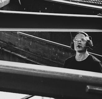 A man wearing protective eyewear and earmuffs appears to be working or observing within an industrial or construction site. Steel beams and metal structures dominate the scene, creating an atmosphere of heavy machinery and engineering.