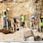 Workers wearing safety gear are engaged in construction activities at a site with rocky terrain. They are handling large metal rods, possibly for reinforcing concrete structures. Piles of earth and construction materials surround the area.