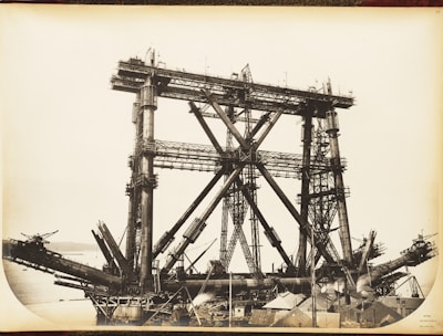 A large, complex metal structure, possibly part of a bridge under construction, dominates the scene. The framework consists of massive steel beams and girders arranged in a triangular pattern. The background includes a body of water and distant hills, while the foreground features various construction materials and small buildings.