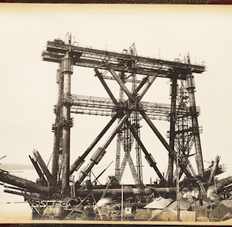 A large, complex metal structure, possibly part of a bridge under construction, dominates the scene. The framework consists of massive steel beams and girders arranged in a triangular pattern. The background includes a body of water and distant hills, while the foreground features various construction materials and small buildings.