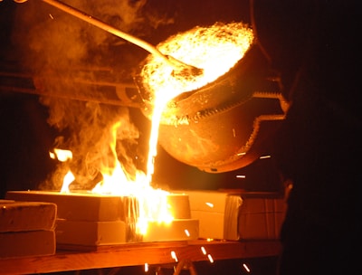 Molten metal is being poured from a large ladle into molds, creating bright orange and yellow flames. Sparks fly as the glowing liquid metal cascades downward, illuminating the surrounding area with intense light.