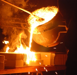 Molten metal is being poured from a large ladle into molds, creating bright orange and yellow flames. Sparks fly as the glowing liquid metal cascades downward, illuminating the surrounding area with intense light.