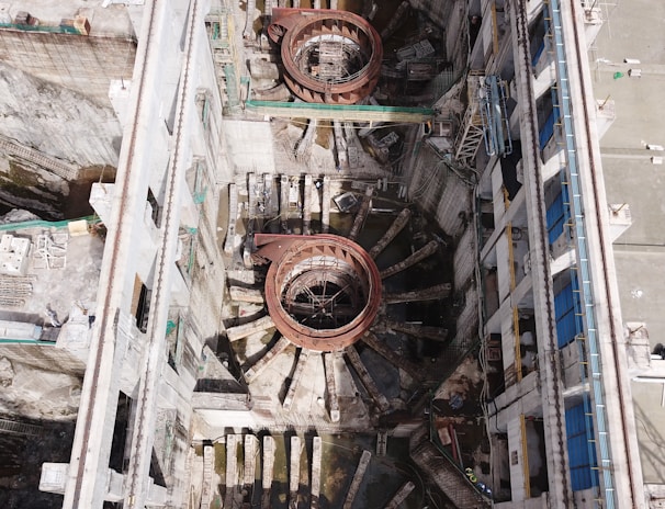 An aerial view of a large industrial or construction site with prominent circular structures and various building materials scattered around. The site is bordered by walls and encompasses machinery designed for heavy-duty work. Details include metallic and concrete textures, with a network of platforms and scaffolding indicating ongoing construction or maintenance activity.