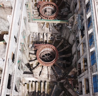 An aerial view of a large industrial or construction site with prominent circular structures and various building materials scattered around. The site is bordered by walls and encompasses machinery designed for heavy-duty work. Details include metallic and concrete textures, with a network of platforms and scaffolding indicating ongoing construction or maintenance activity.