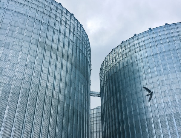 Two large industrial silos dominate the scene, constructed from metallic materials with a corrugated appearance. A yellow Komatsu excavator is visible in the lower foreground, along with a person who appears to be working near the base of one of the silos. A bird can be seen flying in the sky above.