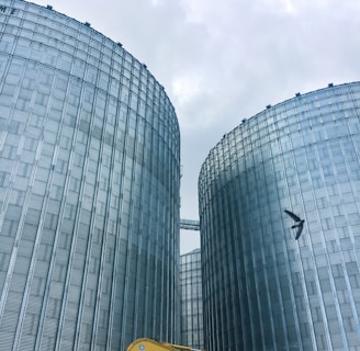 Two large industrial silos dominate the scene, constructed from metallic materials with a corrugated appearance. A yellow Komatsu excavator is visible in the lower foreground, along with a person who appears to be working near the base of one of the silos. A bird can be seen flying in the sky above.