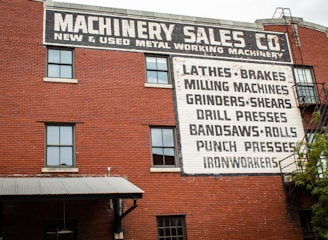 A brick building features multiple windows and a large painted sign advertising a machinery sales company. The sign lists various types of metalworking machinery, including lathes, brakes, milling machines, and more. An external metal staircase runs along the side of the building with some greenery visible at the bottom right.