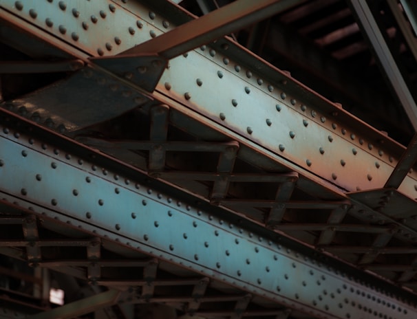 A close-up view of industrial steel girders and beams with a riveted design, predominantly colored in steel blue and rusty orange shades. The intersecting lines and angles create a complex structure, showcasing the metallic texture and utilitarian design.