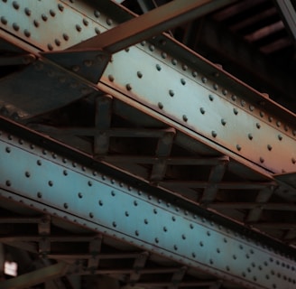 A close-up view of industrial steel girders and beams with a riveted design, predominantly colored in steel blue and rusty orange shades. The intersecting lines and angles create a complex structure, showcasing the metallic texture and utilitarian design.