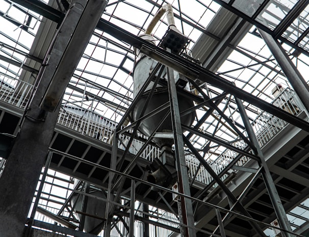 An industrial interior filled with metal beams and supports, showcasing a complex structure of girders and railings. The ceiling is comprised of glass panels allowing natural light to illuminate the space. The architectural design features intricate and heavy-duty construction elements.