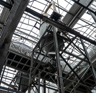 An industrial interior filled with metal beams and supports, showcasing a complex structure of girders and railings. The ceiling is comprised of glass panels allowing natural light to illuminate the space. The architectural design features intricate and heavy-duty construction elements.