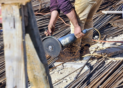 A person is using a power tool to cut through metal rods on a construction site. Sparks are flying from the point of contact between the tool and the metal, and the area is filled with various construction materials like rebar and wooden planks. The person is wearing tan pants and a purple shirt.