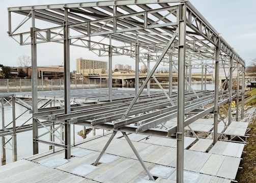 A metal framework structure, likely part of a construction or installation project, is situated near a body of water. The structure comprises numerous horizontal and vertical metal beams, set against an urban backdrop with buildings visible in the distance.