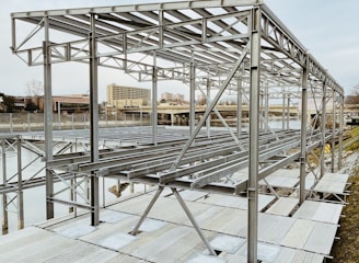 A metal framework structure, likely part of a construction or installation project, is situated near a body of water. The structure comprises numerous horizontal and vertical metal beams, set against an urban backdrop with buildings visible in the distance.