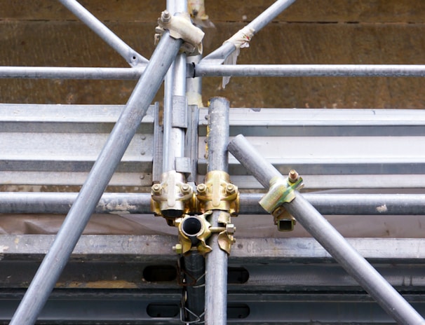 A close-up view of a complex structure made of interlocking metal scaffolding tubes and clamps. The metal is predominantly silver and grey with hints of golden brass at the joints. The scaffolding appears sturdy and is likely used in construction or maintenance work.