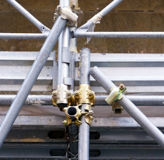 A close-up view of a complex structure made of interlocking metal scaffolding tubes and clamps. The metal is predominantly silver and grey with hints of golden brass at the joints. The scaffolding appears sturdy and is likely used in construction or maintenance work.