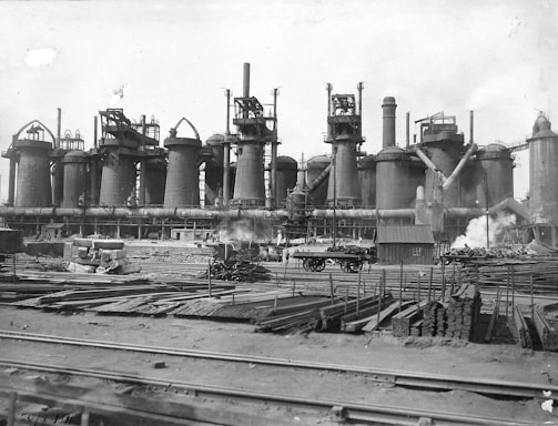 A large industrial facility with multiple tall cylindrical structures, likely blast furnaces, accompanied by numerous pipes and chimneys emitting steam. The foreground contains stacks of metal beams and rails, with scattered construction materials and a railway track.