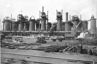 A large industrial facility with multiple tall cylindrical structures, likely blast furnaces, accompanied by numerous pipes and chimneys emitting steam. The foreground contains stacks of metal beams and rails, with scattered construction materials and a railway track.