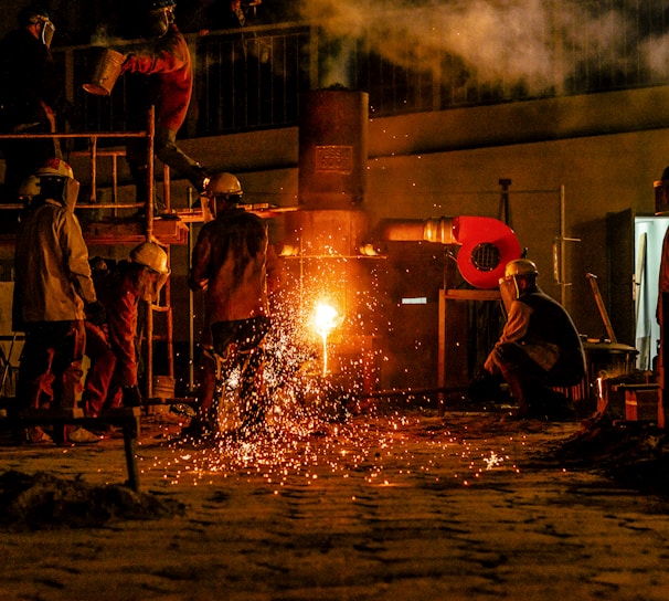 Several workers are wearing protective gear in an industrial setting, engaging in metalworking or a foundry process. Molten metal pours from a furnace, emitting bright sparks and creating a warm glow. Equipment and structural elements surround them, and a haze lingers in the air.