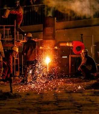 Several workers are wearing protective gear in an industrial setting, engaging in metalworking or a foundry process. Molten metal pours from a furnace, emitting bright sparks and creating a warm glow. Equipment and structural elements surround them, and a haze lingers in the air.