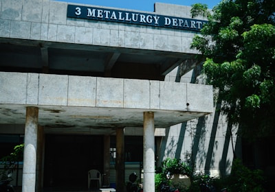 A concrete building with a sign labeled '3 Metallurgy Depart' is partially shaded by a large tree with green foliage. The building has columns supporting a roof over a small entrance area where several motorcycles are parked. The sky is clear and blue, contributing to a sunny atmosphere.