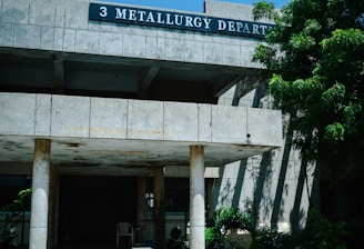 A concrete building with a sign labeled '3 Metallurgy Depart' is partially shaded by a large tree with green foliage. The building has columns supporting a roof over a small entrance area where several motorcycles are parked. The sky is clear and blue, contributing to a sunny atmosphere.