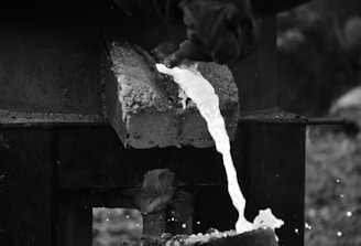 Molten metal is being poured from a furnace. A worker's gloved hand holds the handle of a container, directing the bright, flowing liquid into a mold below. The surrounding environment is industrial, with a focus on the intense heat and fluid motion of the metal.