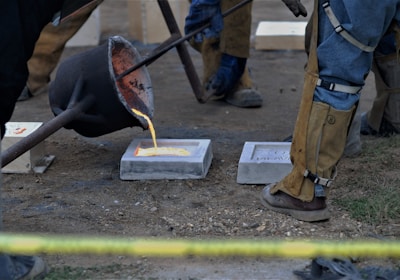 Molten metal is being poured from a crucible into a mold situated on the ground. Several individuals in protective clothing, including heavy-duty boots, leggings, and aprons, are overseeing and facilitating the process. The scene appears to be outdoors, with a visible patch of grass and dirt.